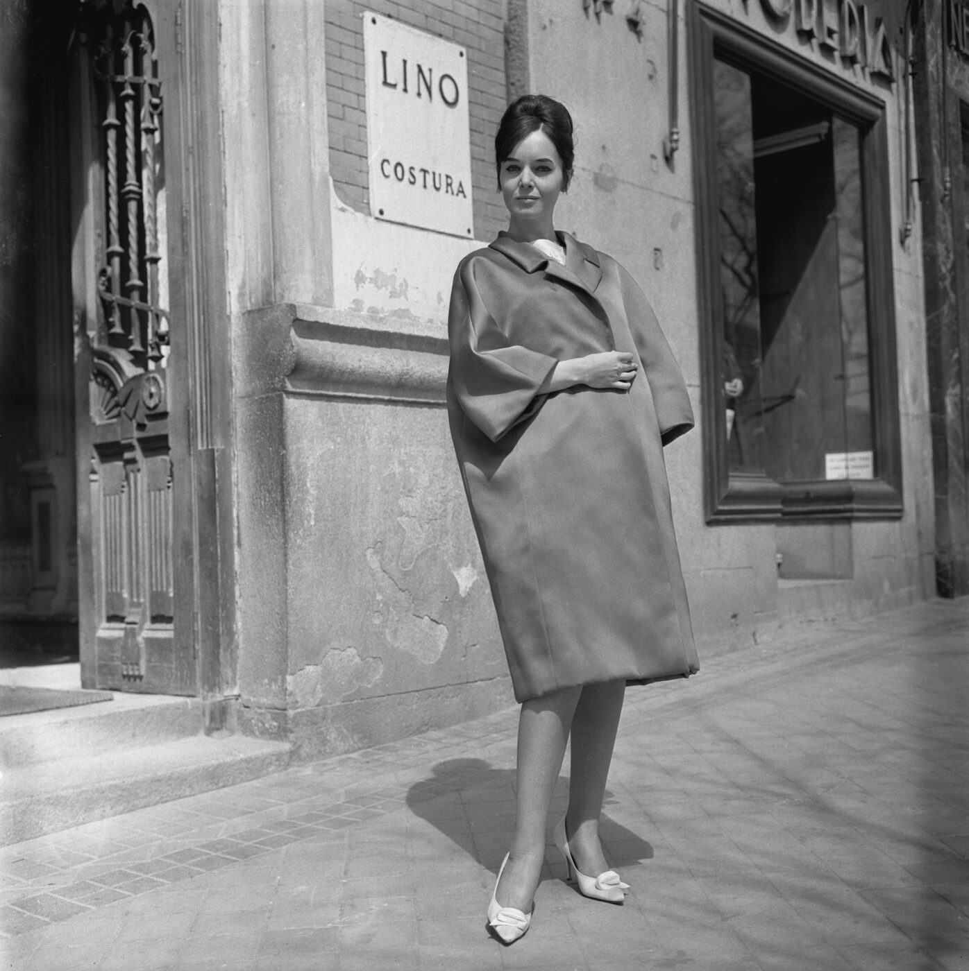 Photographic Social Vision - Maniquí con un modelo del modista Lino (Lino Martínez) enfrente a la entrada de su atelier en la plaza de Santa Bárbara, Madrid, 1962