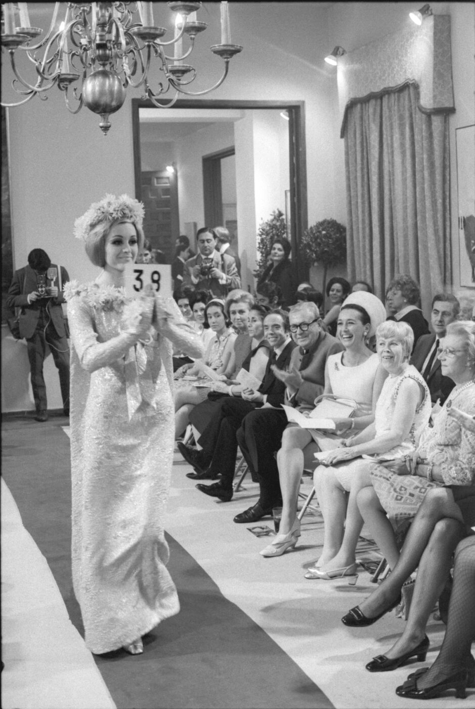 Photographic Social Vision - Desfile de alta costura española en la embajada de los Estados Unidos, Manuel Pertegaz y su sobrina Sionín Caus, Stella Hanania, encargada de compras de los grandes almacenes norteamericanos I. Magnin, la encargada de Bergdof Goodman, junto a Aline Griffith, condesa de Romanones, entre el público asistente, Madrid, enero 1968